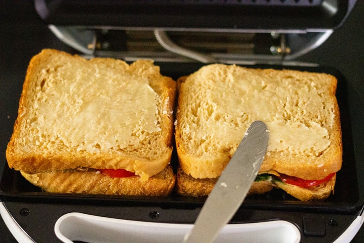 sandwich placed in a grill and butter being spread on top of bread slice