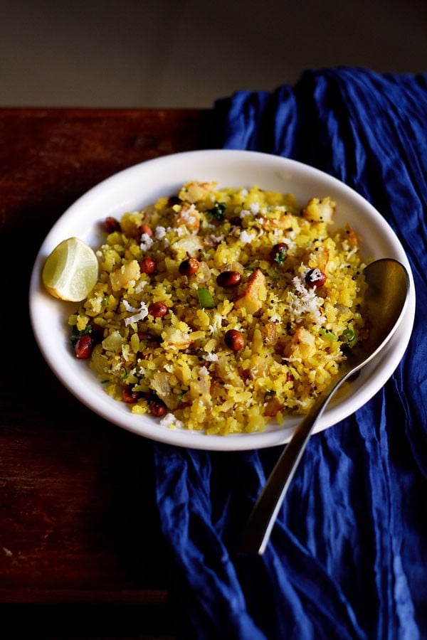 kanda batata poha in a white bowl with a side of lemon wedge and steel spoon on a dark blue fabric