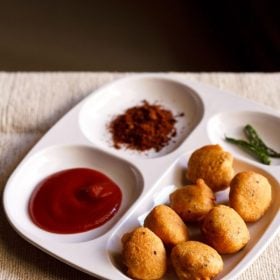 moong dal pakoda served in a white platter with tomato ketchup, dry garlic chutney and fried green chilies.