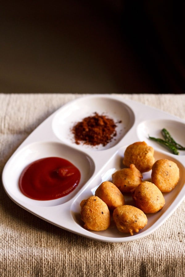 moong dal pakoda served in a white platter with tomato ketchup, dry garlic chutney and fried green chilies.