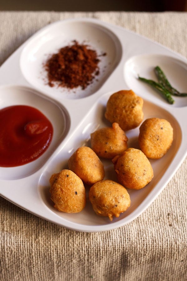 moong dal pakoda served in a white platter with tomato ketchup, dry garlic chutney and fried green chilies. 