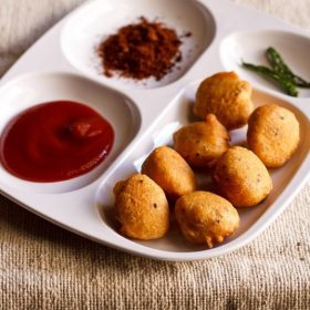 moong dal pakoda served in a white platter with tomato ketchup, dry garlic chutney and fried green chilies.