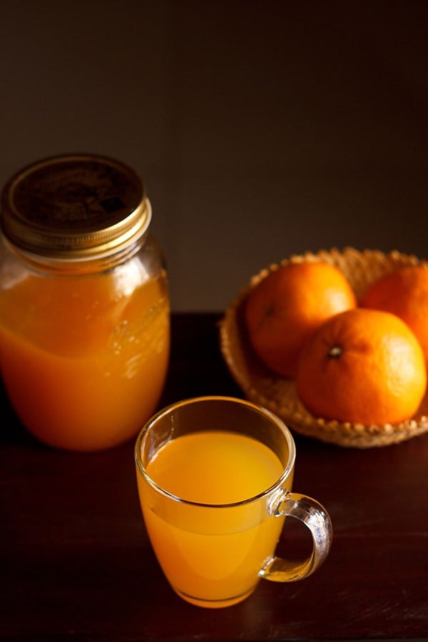 orange squash served in a glass with fresh oranges in a basket and squash in a jug kept at the back.