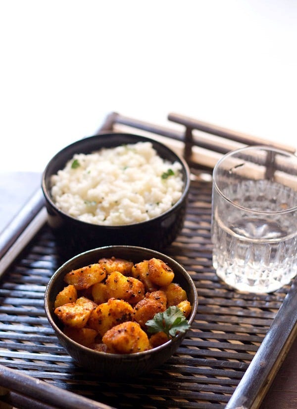 potato roast garnished with a coriander leaf served in a bowl with a bowl of curd rice kept on the left side and a glass of water kept on the right side. 