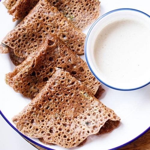 ragi dosa folded and served on a blue rimmed plate with a small blue rimmed bowl of coconut chutney kept on the side.