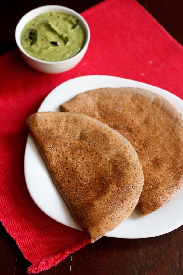 ragi dosa served on a white square plate on a red napkin with a side white bowl of green coconut chutney