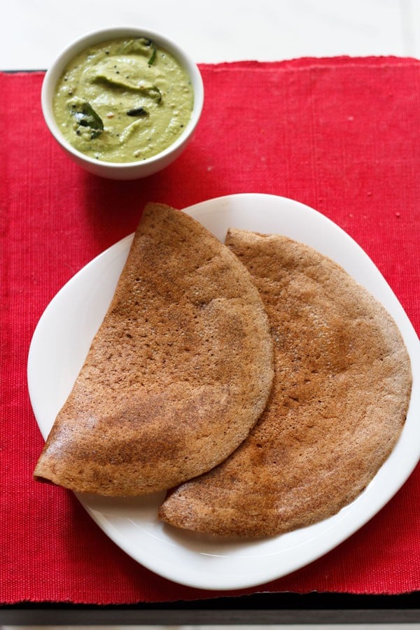 ragi dosa served on a white square plate on a red napkin with a side white bowl of green coconut chutney
