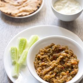 vangyache bharit served in a white bowl placed on a white plate with cucumber fingers kept on the left side, a bowl of curd kept on the top right side and a plate of chapattis kept on the top left side in the background.