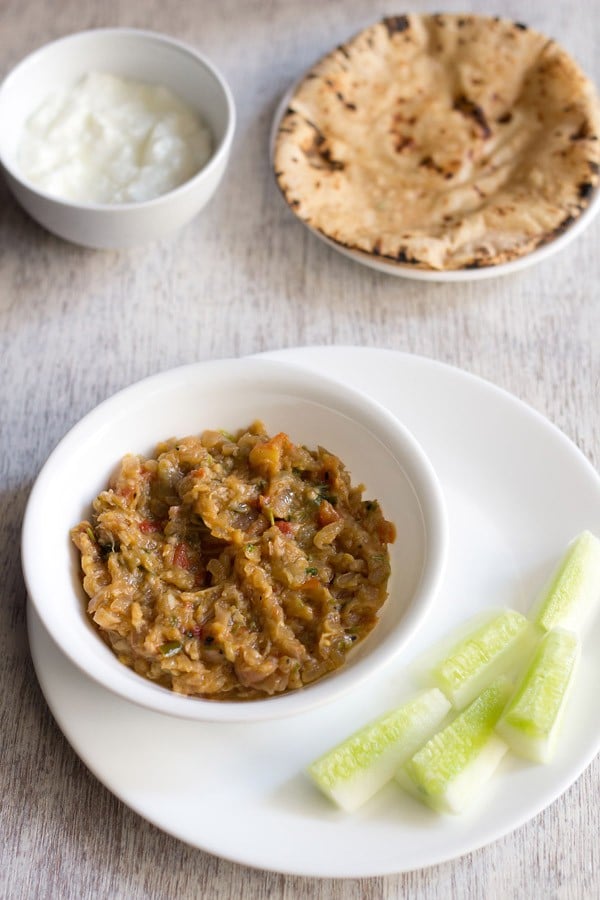 vangyache bharit served in a white bowl placed on a white plate with cucumber fingers kept on the right side, a bowl of curd kept on the top left side and a plate of chapattis kept on the top right side in the background. 