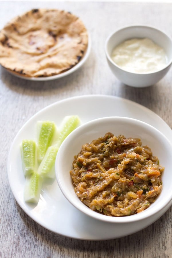 vangyache bharit served in a white bowl placed on a white plate with cucumber fingers kept on the left side, a bowl of curd kept on the top right side and a plate of chapattis kept on the top left side in the background.