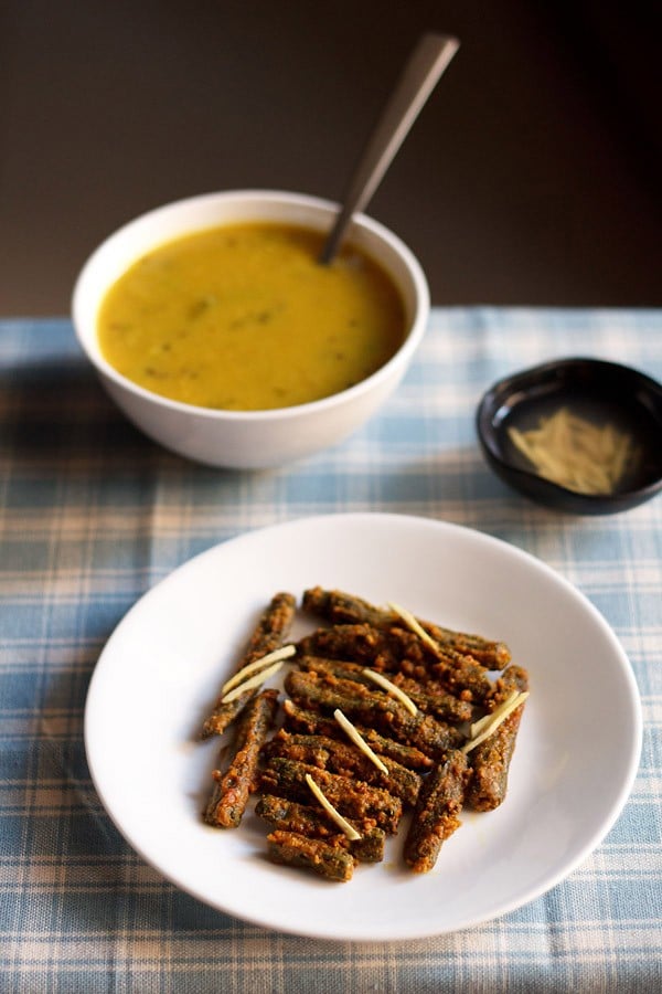 besan bhindi served on a white plate with a bowl of dal and a small bowl of ginger juliennes kept in the background.