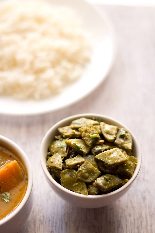 papdi bhaji or sem sabji served in a white bowl with a plate of steamed rice kept in the background and a bowl of sambar kept in the left side. 