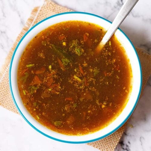 rasam served with a steel spoon in a blue rimmed white bowl placed on a jute mat