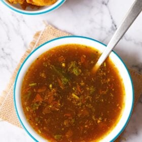 rasam served with a steel spoon in a blue rimmed white bowl placed on a jute mat