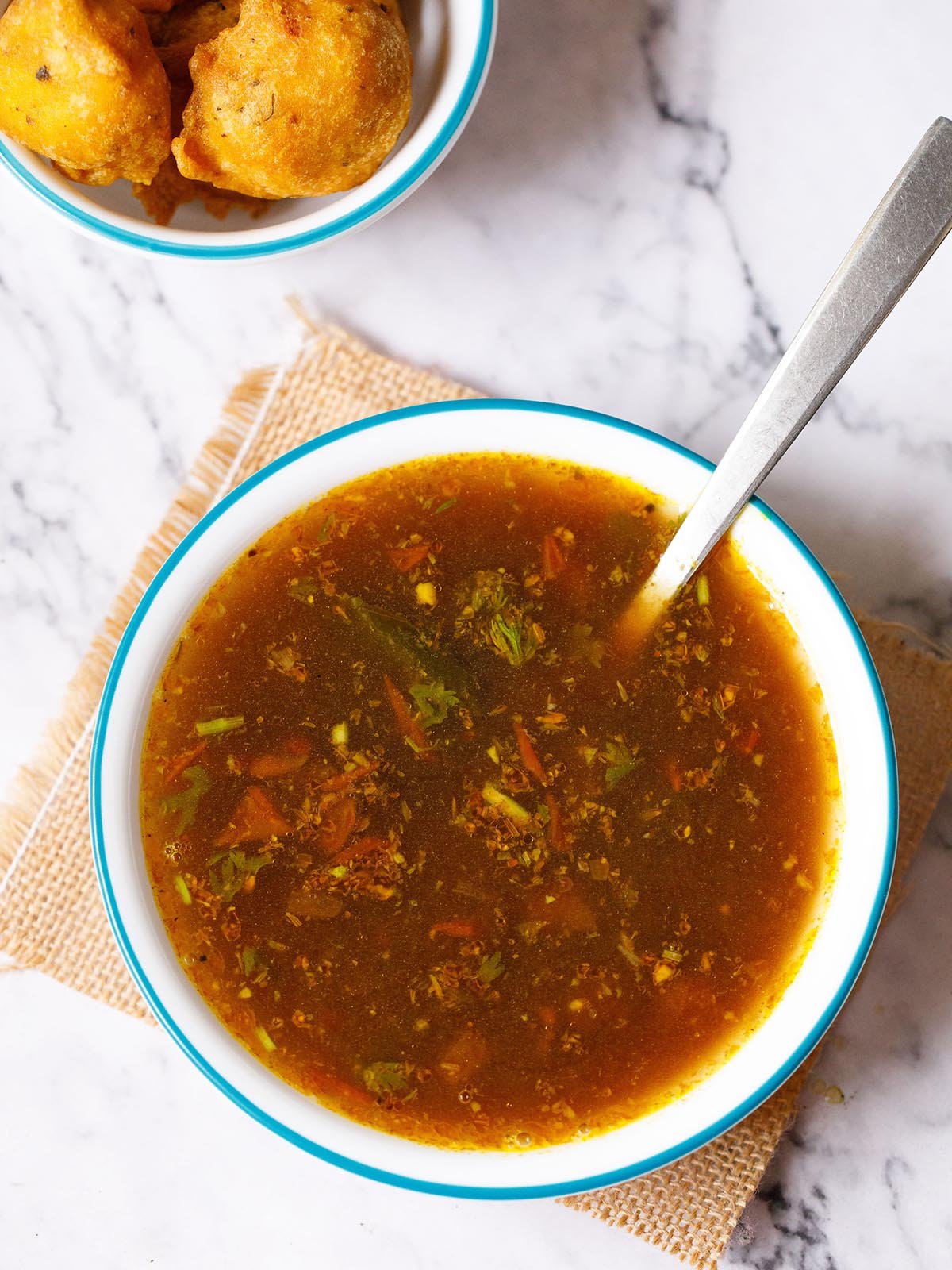 rasam served with a steel spoon in a blue rimmed white bowl placed on a jute mat