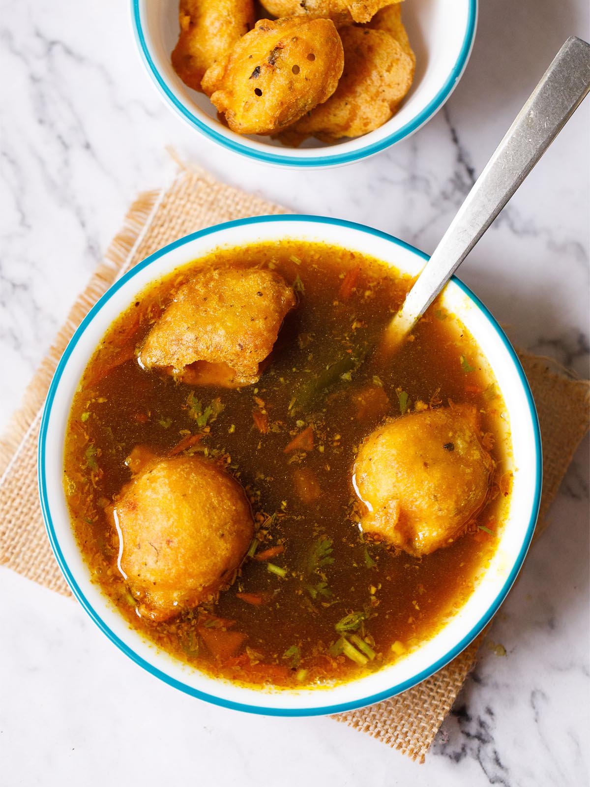 urad dal vada dunked in rasam with a steel spoon served in a blue rimmed white bowl placed on a jute mat