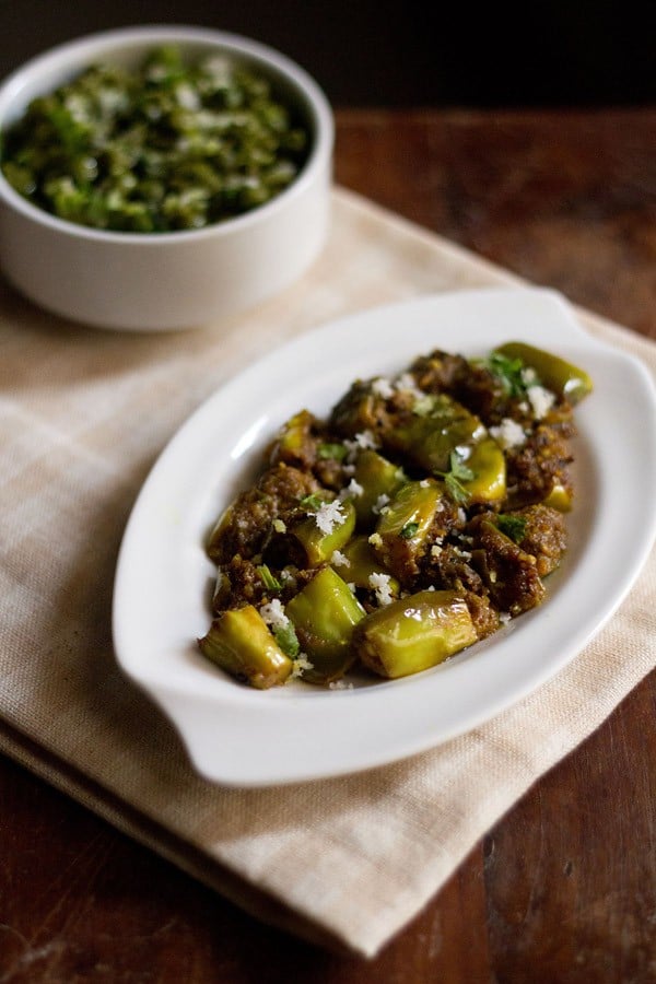 vangyachi bhaji garnished with chopped coriander leaves and fresh grated coconut and served on a white platter with a bowl of sabzi kept in the background.