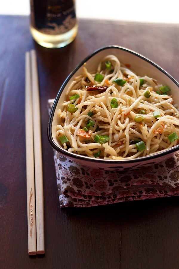 hakka noodles garnished with some spring onion greens and served in a triangular bowl on a printed napkin with cream colored bamboo chopsticks placed at the left side