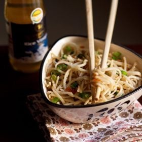 hakka noodles served in a triangular bowl on a printed napkin with cream colored bamboo chopsticks placed on top of the bowl with some hakka noodles between them
