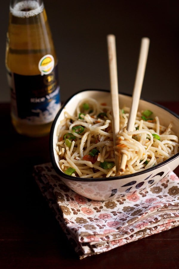 hakka noodles served in a triangular bowl on a printed napkin with cream colored bamboo chopsticks placed on top of the bowl with some hakka noodles between them