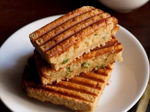 aloo masala ggrilled sandwich served on a white plate with a bowl of tomato ketchup kept on the top right side.