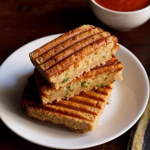 aloo masala ggrilled sandwich served on a white plate with a bowl of tomato ketchup kept on the top right side.