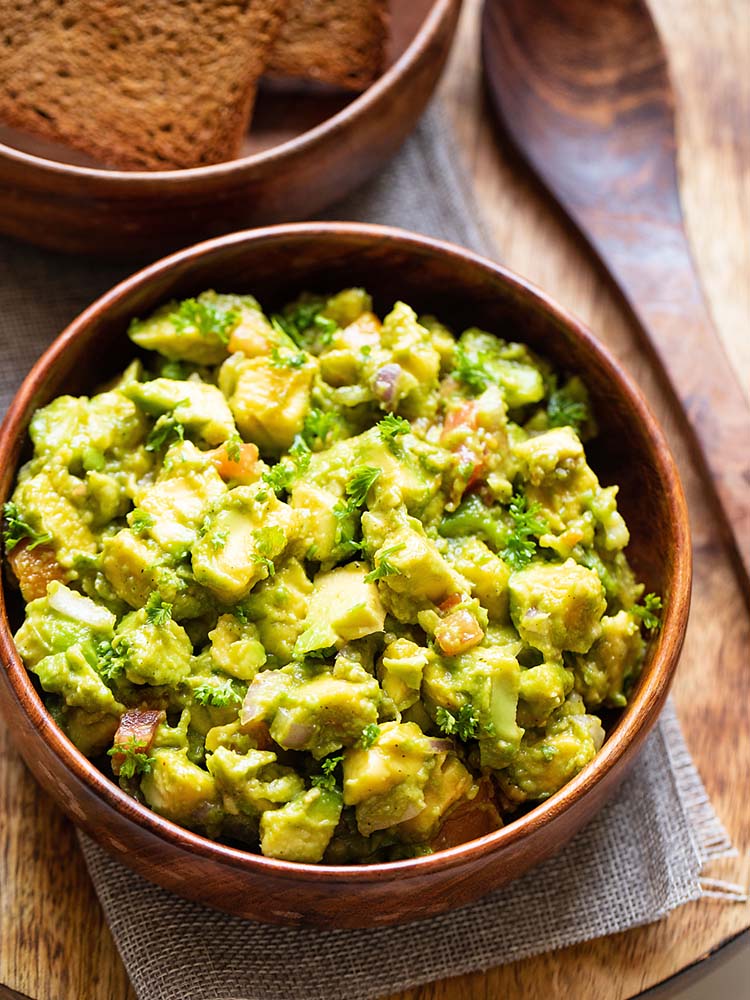 avocado salad served in a wooden bowl with a wooden spoon and some toasted bread in the background.