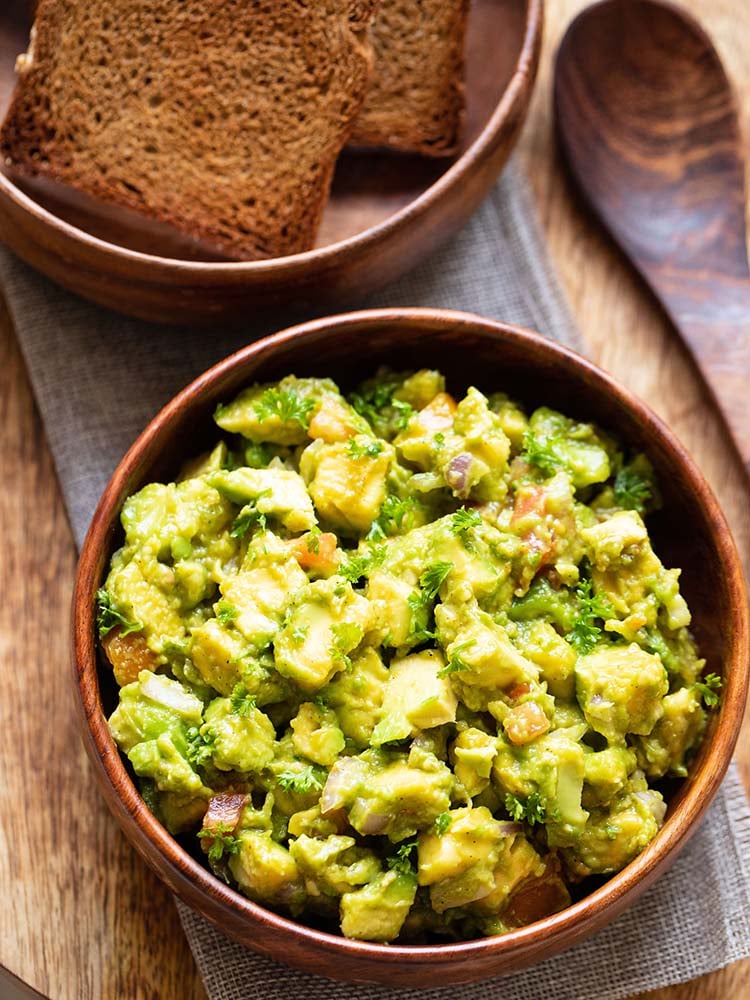 Avocado salad is served in a wooden bowl with a wooden spoon and some toasted bread in the background.