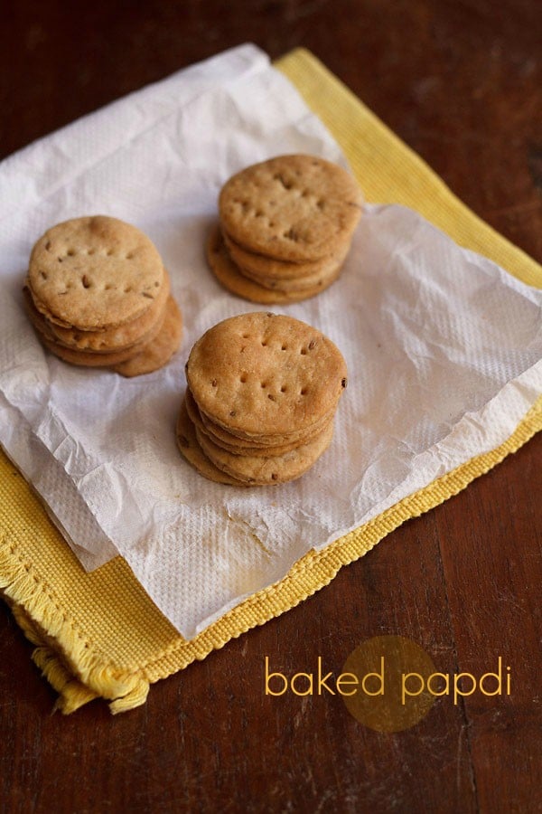 baked papdi stacks placed on a kitchen paper tissue kept on a yellow colored kitchen napkin on a wooden surface with text layover. 