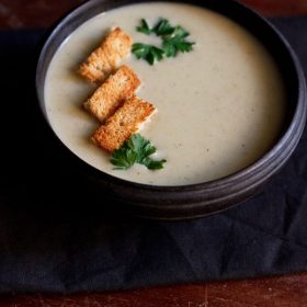 cream of celery soup in a black bowl topped with bread croutons and a parsley sprig.