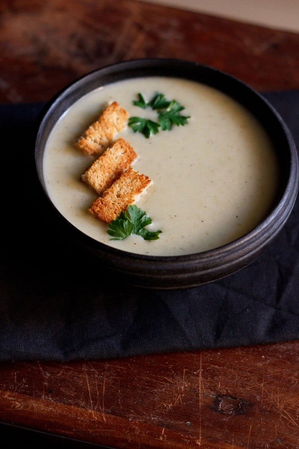 cream of celery soup in a black bowl topped with bread croutons and a parsley sprig.