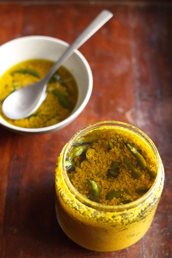hari mirch ka achar in glass jar with a bowl filled with the chilli pickle and a spoon on top side.