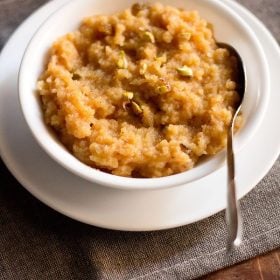 moong dal halwa served in a white bowl on a white plate with a spoon inside the bowl.