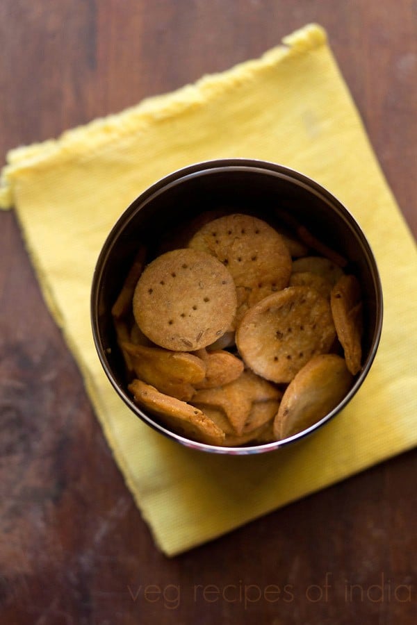 papdi placed in a steel box kept on a yellow colored kitchen napkin on a wooden surface with text layover. 