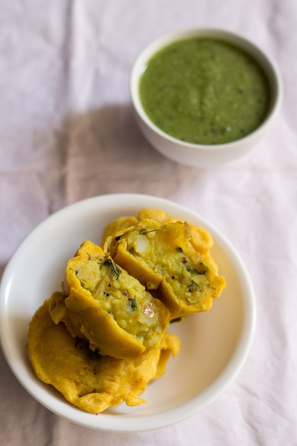 aloo bonda halved and served on a whole aloo bonda on a white plate with a small bowl of green chutney kept on the top right side. 