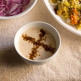 burani raita in a white bowl with vegetable biryani and some sliced red onions on a cream jute napkin