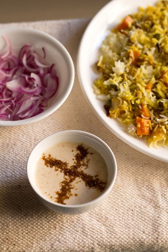 burani raita sprinkled with roasted cumin powder and served in a small bowl with a plate of veg biryani and a bowl of onion slices in the background. 