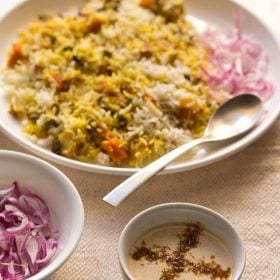 burani raita in a white bowl with vegetable biryani and some sliced red onions on a cream jute napkin