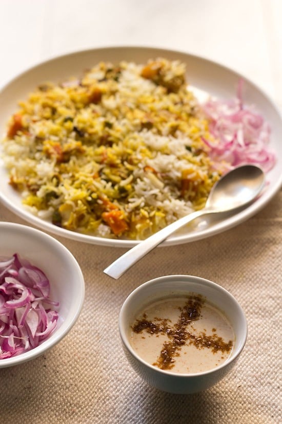 burani raita in a white bowl with vegetable biryani and some sliced red onions on a cream jute napkin