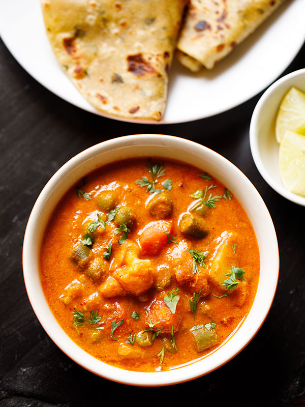 instant pot korma served in an off-white bowl with a plate of indian flatbreads and a bowl of chopped lemons on a black slate board