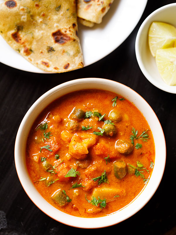 instant pot veg korma served in an off-white bowl with a plate of Indian flatbreads and a bowl of chopped lemons on a black slate board