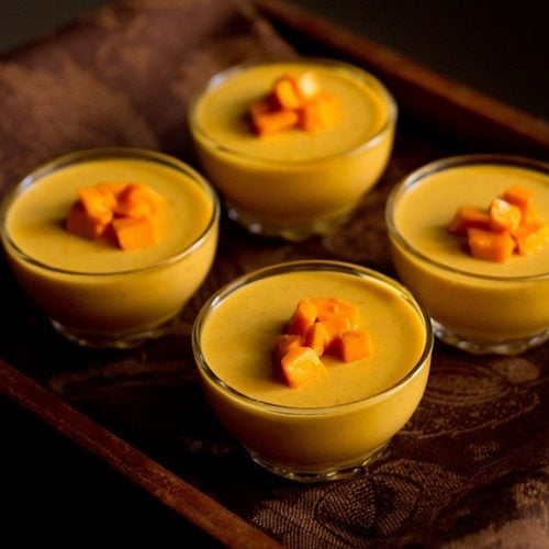 four bowls of vegetarian mango panna cotta topped with fresh cubes of mango on a wooden serving platter