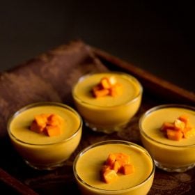 four bowls of vegetarian mango panna cotta topped with fresh cubes of mango on a wooden serving platter.