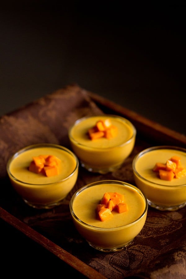 four bowls of vegetarian mango panna cotta topped with fresh cubes of mango on a wooden serving platter.