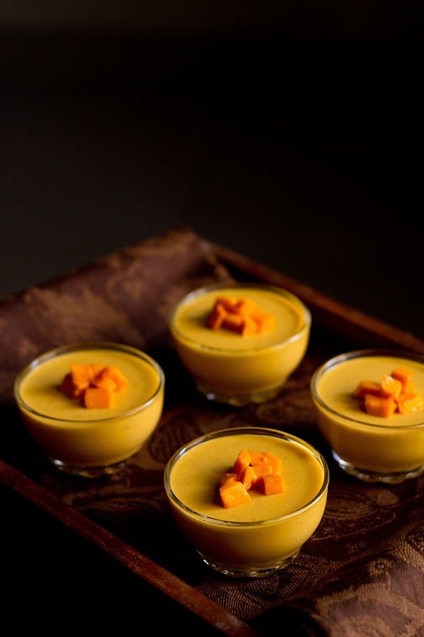 four bowls of vegetarian mango panna cotta topped with fresh cubes of mango on a wooden serving platter.