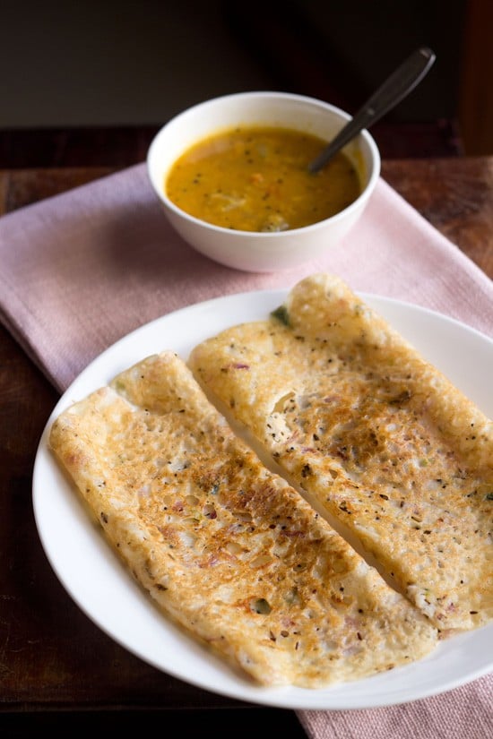 Onion rava dosa is served on a white plate alongside sambar.