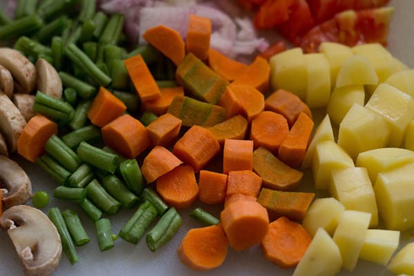 chopped carrots, green beans, potatoes, onions, tomatoes and button mushrooms on a chopping board