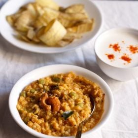 bisi bele bath served in a white bowl with a spoon in it and a bowl of banana chips and small bowl of raita kept in top left and right side.