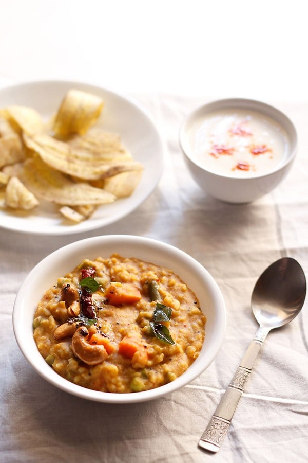 bisi bele bath served in a white bowl with a spoon kept on the right side,  bowl of banana chips kept on the top left side and a small bowl of raita kept on the top right side.