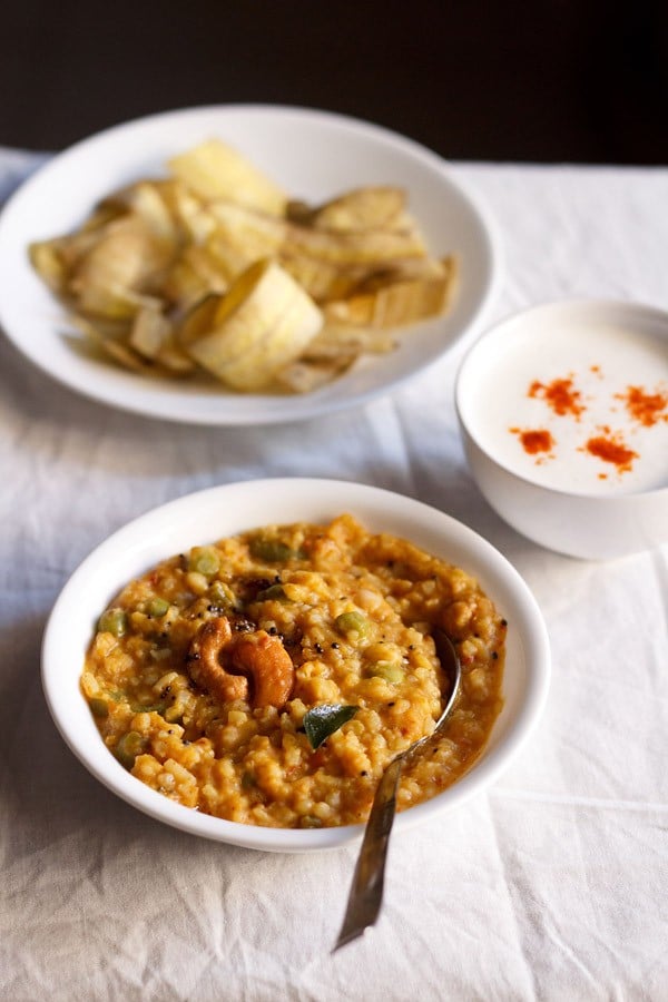 bisi bele bath served in a white bowl with a spoon in it and a bowl of banana chips and small bowl of raita kept in top left and right side.